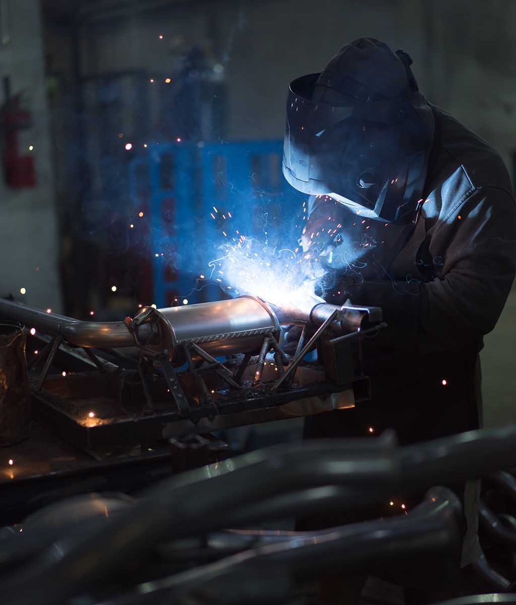 dramatic view of a worker wearing protective gear, welding together pieces of an exhaust pipe, with several other pipes around, on the work table, and metal blue frames in the background, in an industrial setting